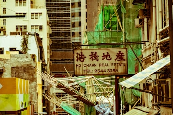 A cityscape with a real estate sign in the foreground, prominently displaying the name and phone number. Surrounding the sign are various buildings with different architectural styles, some with scaffolding and green netting. The scene features a mix of older, worn structures alongside modern glass-fronted buildings. Power lines and construction materials add to the urban complexity.
