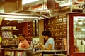 A traditional apothecary setting with two people engaged in discussion or transaction. Wooden drawers line the wall, indicating storage for herbs or medicines. The environment is warm and reflects the style of an old-world herbal medicine shop.