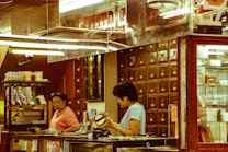 A traditional apothecary setting with two people engaged in discussion or transaction. Wooden drawers line the wall, indicating storage for herbs or medicines. The environment is warm and reflects the style of an old-world herbal medicine shop.
