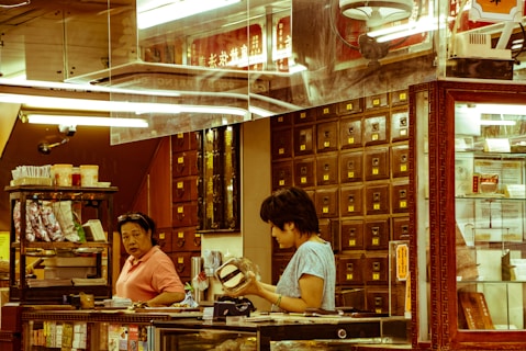A traditional apothecary setting with two people engaged in discussion or transaction. Wooden drawers line the wall, indicating storage for herbs or medicines. The environment is warm and reflects the style of an old-world herbal medicine shop.