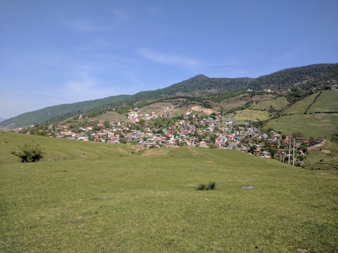 A small village nestled in a lush valley surrounded by rolling hills and dense forests. The village is made up of numerous colorful houses clustered together, with winding paths and roads connecting them. The foreground features a wide expanse of green grass under a clear blue sky.