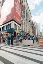 A bustling city street scene featuring a large department store with a prominent red sign. Pedestrians walk along the sidewalk, some of whom are interacting near food carts and street vendors. The building's sign reads 'The World's Largest Store' with additional branding visible. American flags are displayed on the building, and the scene conveys busy urban life.