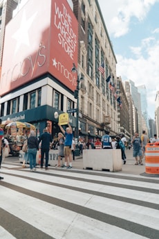 A bustling city street scene featuring a large department store with a prominent red sign. Pedestrians walk along the sidewalk, some of whom are interacting near food carts and street vendors. The building's sign reads 'The World's Largest Store' with additional branding visible. American flags are displayed on the building, and the scene conveys busy urban life.