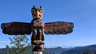 A totem pole featuring a stylized bird with outspread wings is prominently displayed against a clear blue sky. The pole exhibits intricate carvings with vibrant colors including red, black, and natural wood tones. A forested landscape with distant hills and water is visible in the background.