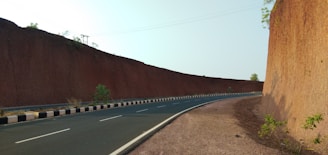 A winding road between two high earthen walls, bordered by sparse vegetation and marked by a clear blue sky above. The road is paved and has white lane markings, with a black and white striped barrier on one side.