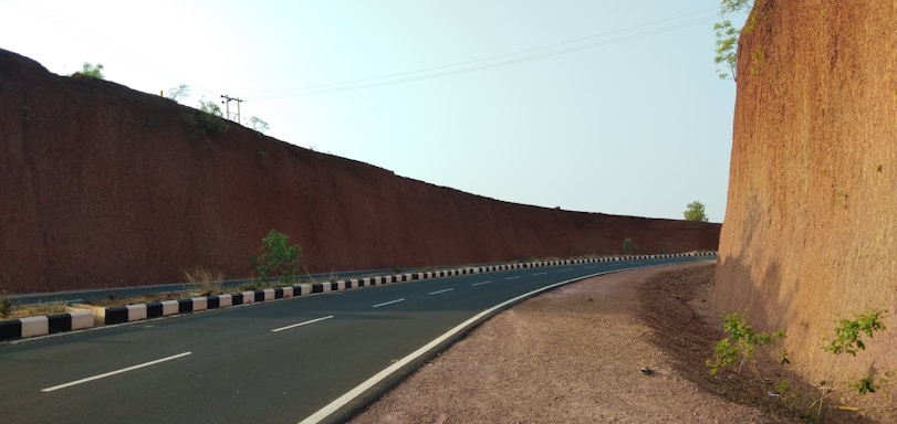 A winding road between two high earthen walls, bordered by sparse vegetation and marked by a clear blue sky above. The road is paved and has white lane markings, with a black and white striped barrier on one side.
