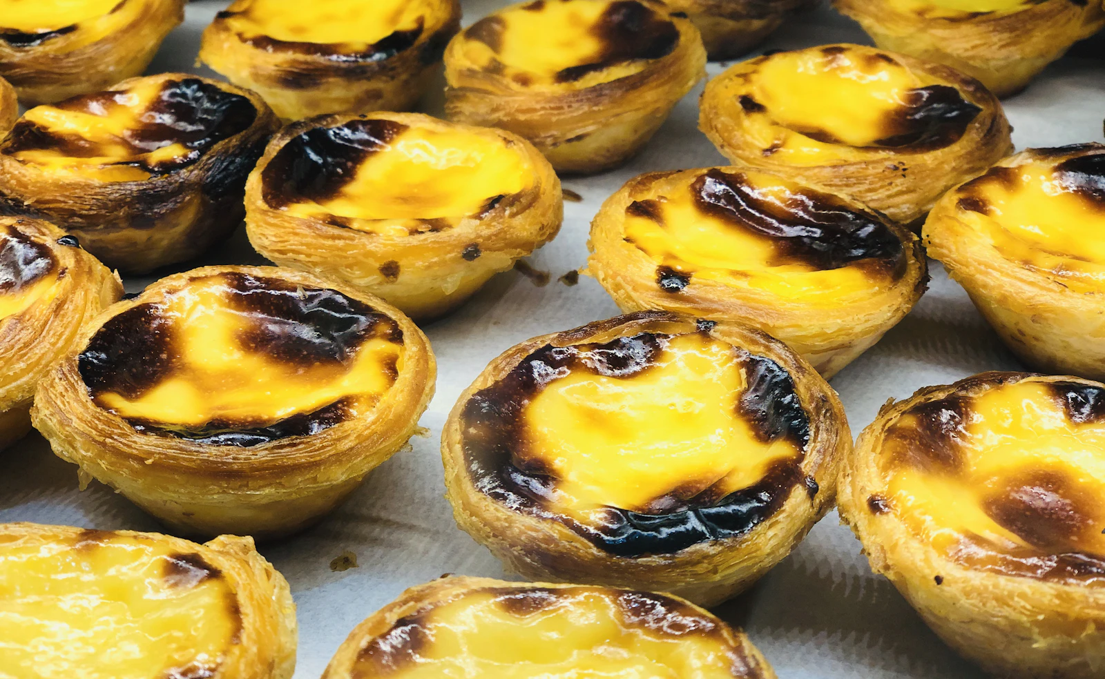 Rows of freshly baked pasteis de nata with flaky golden pastry shells and caramelised, blistered custard tops