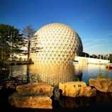 A large geodesic dome with a triangular grid pattern stands by a tranquil body of water, reflecting in the calm surface. Surrounding the dome are lush trees and a clear blue sky, contributing to a serene and peaceful atmosphere. Large rocks are situated in the foreground near the water's edge.