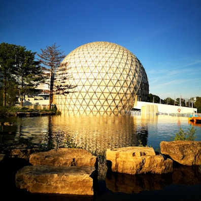 A large geodesic dome with a triangular grid pattern stands by a tranquil body of water, reflecting in the calm surface. Surrounding the dome are lush trees and a clear blue sky, contributing to a serene and peaceful atmosphere. Large rocks are situated in the foreground near the water's edge.