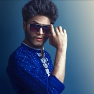 A person wearing a deep blue traditional outfit with patterned details holds sunglasses with one hand against a gradient blue background. The hair is styled with volume, and the pose conveys confidence and a playful attitude.