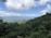 Dense, lush green tropical rainforest extends to the horizon under a blue sky with scattered clouds. Rolling hills and distant mountains are visible in the background, creating a sense of vastness and natural beauty.