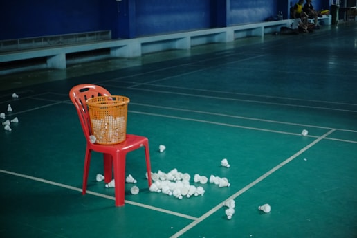 A red plastic chair is positioned on a green badminton court. A yellow basket filled with shuttlecocks is placed on the chair, while more shuttlecocks are scattered on the floor around it. The background features a bench against a dark blue wall, with a few people sitting in the far corner.