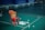 A red plastic chair is positioned on a green badminton court. A yellow basket filled with shuttlecocks is placed on the chair, while more shuttlecocks are scattered on the floor around it. The background features a bench against a dark blue wall, with a few people sitting in the far corner.