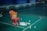 A red plastic chair is positioned on a green badminton court. A yellow basket filled with shuttlecocks is placed on the chair, while more shuttlecocks are scattered on the floor around it. The background features a bench against a dark blue wall, with a few people sitting in the far corner.