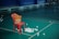 A red plastic chair is positioned on a green badminton court. A yellow basket filled with shuttlecocks is placed on the chair, while more shuttlecocks are scattered on the floor around it. The background features a bench against a dark blue wall, with a few people sitting in the far corner.