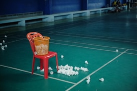 A red plastic chair is positioned on a green badminton court. A yellow basket filled with shuttlecocks is placed on the chair, while more shuttlecocks are scattered on the floor around it. The background features a bench against a dark blue wall, with a few people sitting in the far corner.