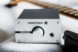 An audio amplifier with a silver front panel labeled 'ERZETICH' and 'Bacillus'. The front features a large volume knob and headphone jack. It is placed on a textured white surface with part of a guitar neck visible in the background, creating a tech and music-focused setting.