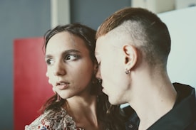 A young woman with long dark hair is being leaned on by a man with short, shaved hair and an earring. The woman appears to be looking away with a thoughtful expression. Both individuals are in close proximity, suggesting intimacy. The background is out of focus with muted colors and a red rectangular object.