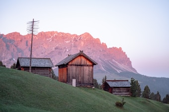 A serene mountain landscape during sunrise or sunset, featuring rustic wooden cabins situated on a grassy hillside. The sky displays a soft pastel gradient, and the mountains in the background are bathed in a gentle pink hue from the light. Sparse vegetation and a tall antenna add to the scenic composition.