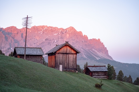 A serene mountain landscape during sunrise or sunset, featuring rustic wooden cabins situated on a grassy hillside. The sky displays a soft pastel gradient, and the mountains in the background are bathed in a gentle pink hue from the light. Sparse vegetation and a tall antenna add to the scenic composition.
