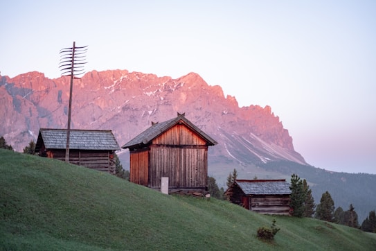 A serene mountain landscape during sunrise or sunset, featuring rustic wooden cabins situated on a grassy hillside. The sky displays a soft pastel gradient, and the mountains in the background are bathed in a gentle pink hue from the light. Sparse vegetation and a tall antenna add to the scenic composition.