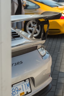 A close-up view of the rear end of a silver sports car, featuring a large spoiler and visible GT3RS branding. The license plate from British Columbia reads 'GB9 11M' with a sticker displaying the date March 2019. In the background, part of a yellow sports car is visible, as well as blurred figures of people.