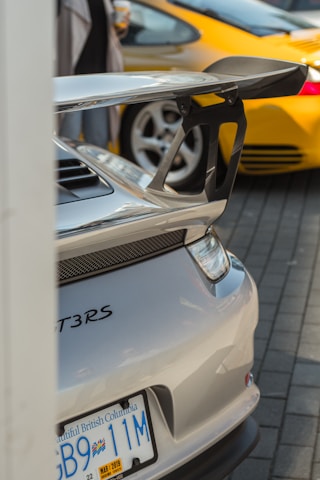 A close-up view of the rear end of a silver sports car, featuring a large spoiler and visible GT3RS branding. The license plate from British Columbia reads 'GB9 11M' with a sticker displaying the date March 2019. In the background, part of a yellow sports car is visible, as well as blurred figures of people.