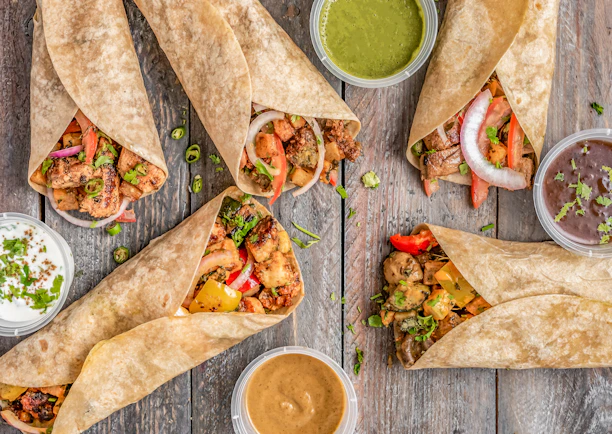 A vibrant spread of colorful Moroccan-style tacos, fresh pizzas, and hearty sandwiches on a rustic wooden table.