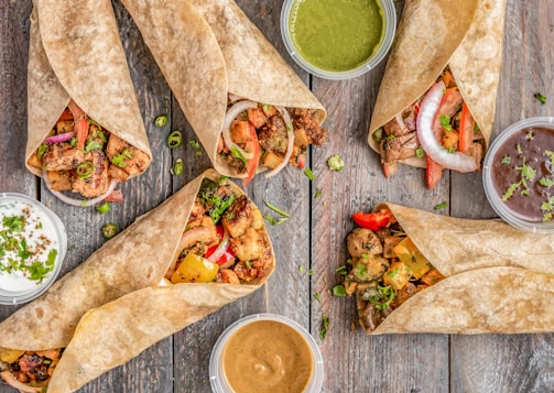 A rustic wooden table serves as the backdrop for a diverse assortment of wraps filled with colorful grilled vegetables, sliced onions, and chunks of seasoned meat or chicken. Small bowls of assorted sauces accompany the wraps, featuring green, brown, and creamy beige hues.