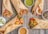 A rustic wooden table serves as the backdrop for a diverse assortment of wraps filled with colorful grilled vegetables, sliced onions, and chunks of seasoned meat or chicken. Small bowls of assorted sauces accompany the wraps, featuring green, brown, and creamy beige hues.