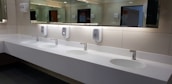 A clean and modern public restroom with a long white countertop featuring four sinks. Each sink is equipped with a chrome faucet and is paired with a hand soap dispenser mounted on a tiled wall. Large mirrors span the length of the countertop, reflecting the restroom's lighting and interior.