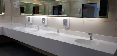 A clean and modern public restroom with a long white countertop featuring four sinks. Each sink is equipped with a chrome faucet and is paired with a hand soap dispenser mounted on a tiled wall. Large mirrors span the length of the countertop, reflecting the restroom's lighting and interior.