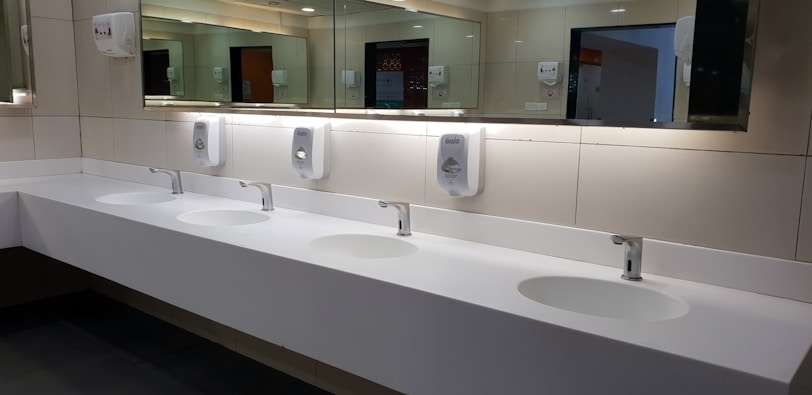 A clean and modern public restroom with a long white countertop featuring four sinks. Each sink is equipped with a chrome faucet and is paired with a hand soap dispenser mounted on a tiled wall. Large mirrors span the length of the countertop, reflecting the restroom's lighting and interior.