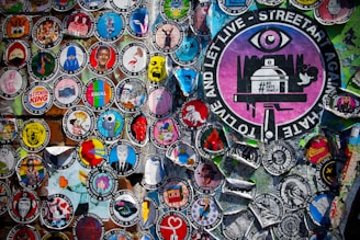 A vibrant collection of circular street art stickers with various illustrations and messages, including a prominent large sticker featuring an eye and the text 'TO LIVE AND LET LIVE - STREETART AGAINST HATE.' The stickers display a wide range of artistic styles and colorful graphics against a textured background.