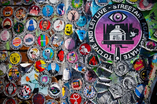 A vibrant collection of circular street art stickers with various illustrations and messages, including a prominent large sticker featuring an eye and the text 'TO LIVE AND LET LIVE - STREETART AGAINST HATE.' The stickers display a wide range of artistic styles and colorful graphics against a textured background.
