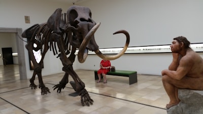 A museum exhibit featuring a large mammoth skeleton on display next to a sculpture of a prehistoric human sitting thoughtfully. A person wearing a red outfit is seated on a bench, observing the display. The room is spacious with beige tiles and minimalistic design.