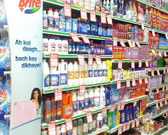 A supermarket aisle filled with various cleaning products, organized neatly on shelves. Promotional sale tags are visible on many items, indicating discounted prices. A promotional stand-up banner on the left features a smiling woman holding clean laundry, with a slogan in Hindi.
