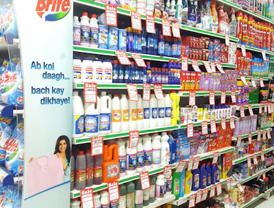 A supermarket aisle filled with various cleaning products, organized neatly on shelves. Promotional sale tags are visible on many items, indicating discounted prices. A promotional stand-up banner on the left features a smiling woman holding clean laundry, with a slogan in Hindi.
