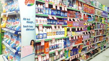 A supermarket aisle filled with various cleaning products, organized neatly on shelves. Promotional sale tags are visible on many items, indicating discounted prices. A promotional stand-up banner on the left features a smiling woman holding clean laundry, with a slogan in Hindi.