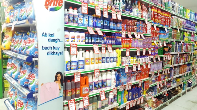A supermarket aisle filled with various cleaning products, organized neatly on shelves. Promotional sale tags are visible on many items, indicating discounted prices. A promotional stand-up banner on the left features a smiling woman holding clean laundry, with a slogan in Hindi.