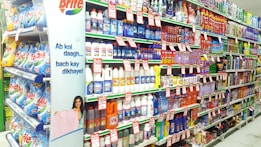 A supermarket aisle filled with various cleaning products, organized neatly on shelves. Promotional sale tags are visible on many items, indicating discounted prices. A promotional stand-up banner on the left features a smiling woman holding clean laundry, with a slogan in Hindi.