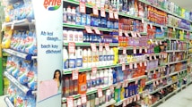 A supermarket aisle filled with various cleaning products, organized neatly on shelves. Promotional sale tags are visible on many items, indicating discounted prices. A promotional stand-up banner on the left features a smiling woman holding clean laundry, with a slogan in Hindi.