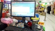 A checkout counter in a grocery store with a computer screen displaying a point-of-sale software interface. The counter has various products like snacks and a barcode scanner. Shelves filled with assorted items are visible in the background, with customers browsing and shopping.