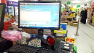 A checkout counter in a grocery store with a computer screen displaying a point-of-sale software interface. The counter has various products like snacks and a barcode scanner. Shelves filled with assorted items are visible in the background, with customers browsing and shopping.