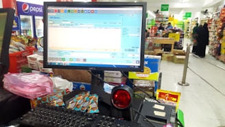 A checkout counter in a grocery store with a computer screen displaying a point-of-sale software interface. The counter has various products like snacks and a barcode scanner. Shelves filled with assorted items are visible in the background, with customers browsing and shopping.