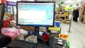 A checkout counter in a grocery store with a computer screen displaying a point-of-sale software interface. The counter has various products like snacks and a barcode scanner. Shelves filled with assorted items are visible in the background, with customers browsing and shopping.