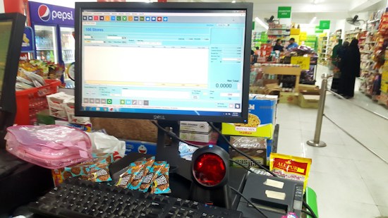 A checkout counter in a grocery store with a computer screen displaying a point-of-sale software interface. The counter has various products like snacks and a barcode scanner. Shelves filled with assorted items are visible in the background, with customers browsing and shopping.