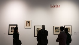 Several people are observing framed artwork on a gallery wall. The artwork consists of portraits and illustrations with a variety of styles. A textual installation on the wall reads 'New [wo]man'. Lighting is subdued, creating an intimate atmosphere.