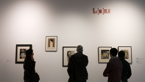 Several people are observing framed artwork on a gallery wall. The artwork consists of portraits and illustrations with a variety of styles. A textual installation on the wall reads 'New [wo]man'. Lighting is subdued, creating an intimate atmosphere.