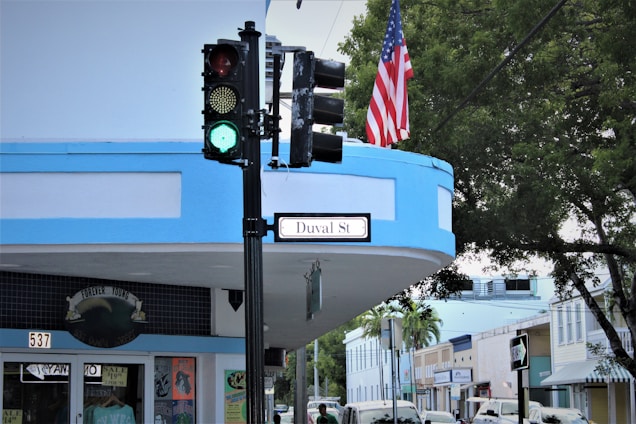 A street corner featuring a green traffic light, a brightly colored storefront with a sign that says 'Duval St', and an American flag. Buildings line the street under a clear sky, with trees providing some greenery.